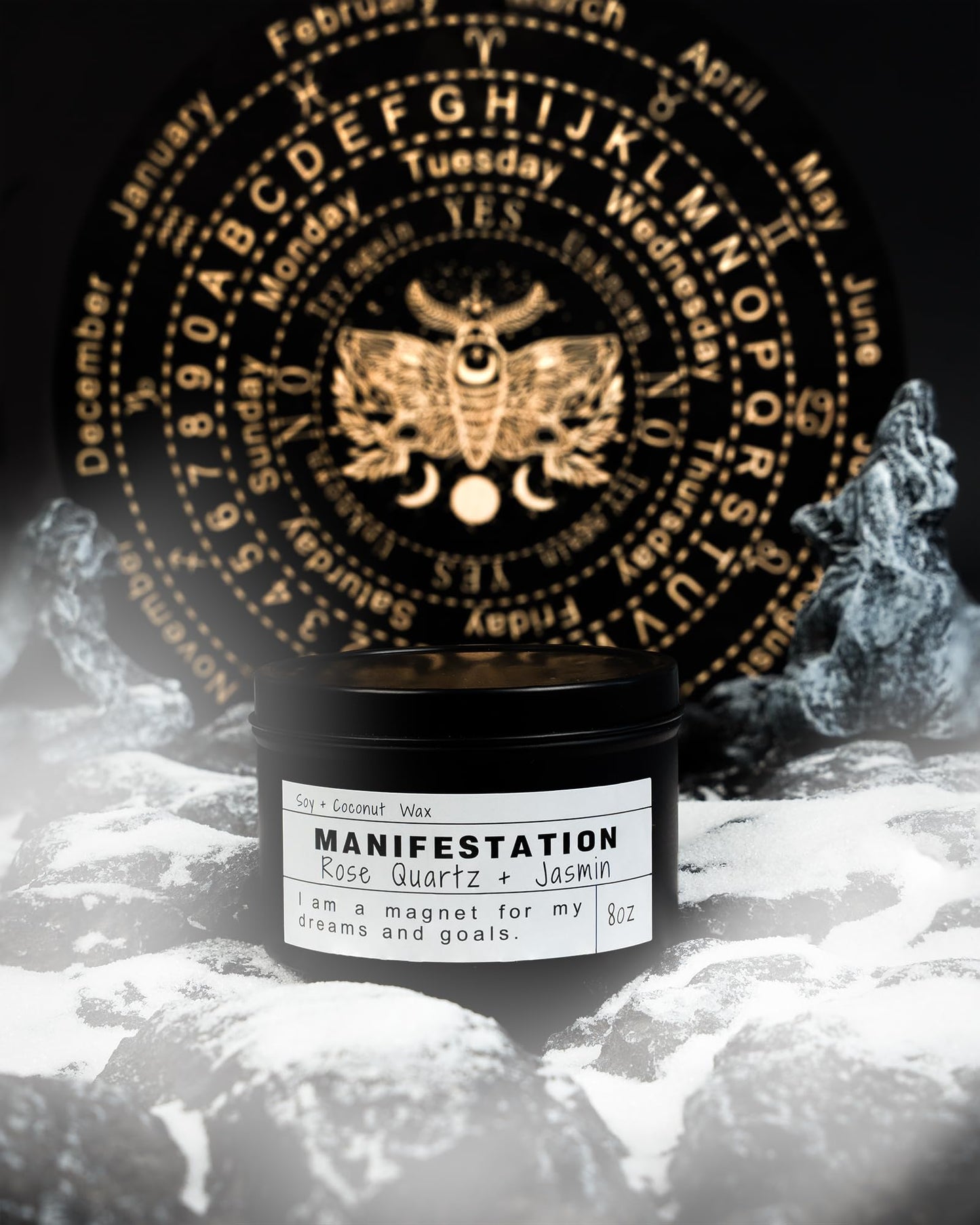 Black Manifestation candle with rose quartz and jasmin label, displayed on snowy rocks with a mystical gold symbol wheel in the background.