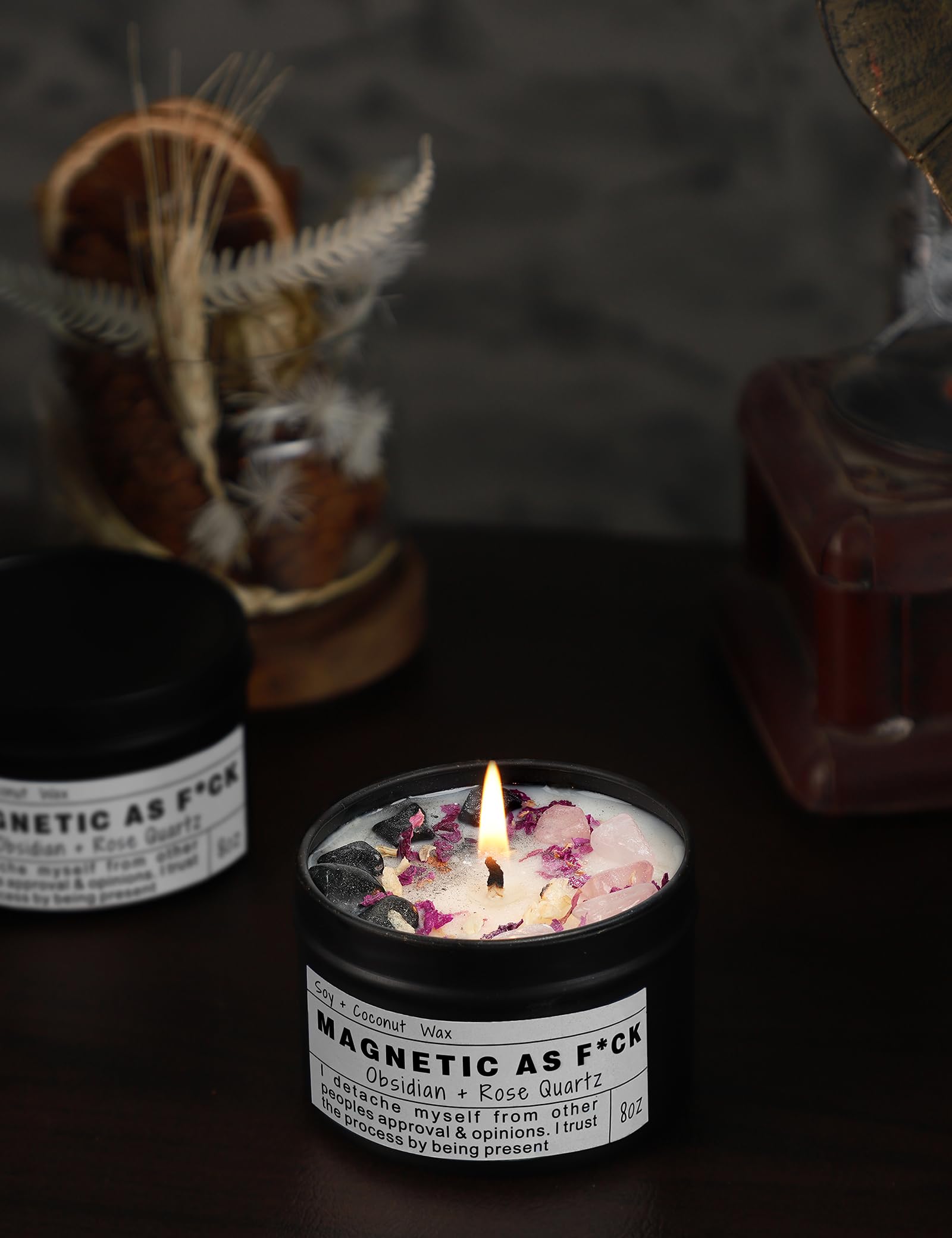 Magnetic As F*ck candle with obsidian and rose quartz crystals burning in a black tin on a dark background.