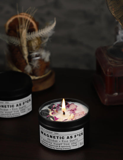 Magnetic As F*ck candle with obsidian and rose quartz crystals burning in a black tin on a dark background.