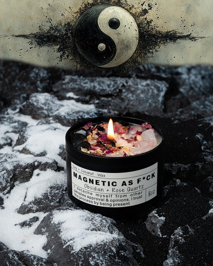 Magnetic As F*ck candle with obsidian and rose quartz crystals burning on a snowy rock surface with a yin-yang illustration in the background.