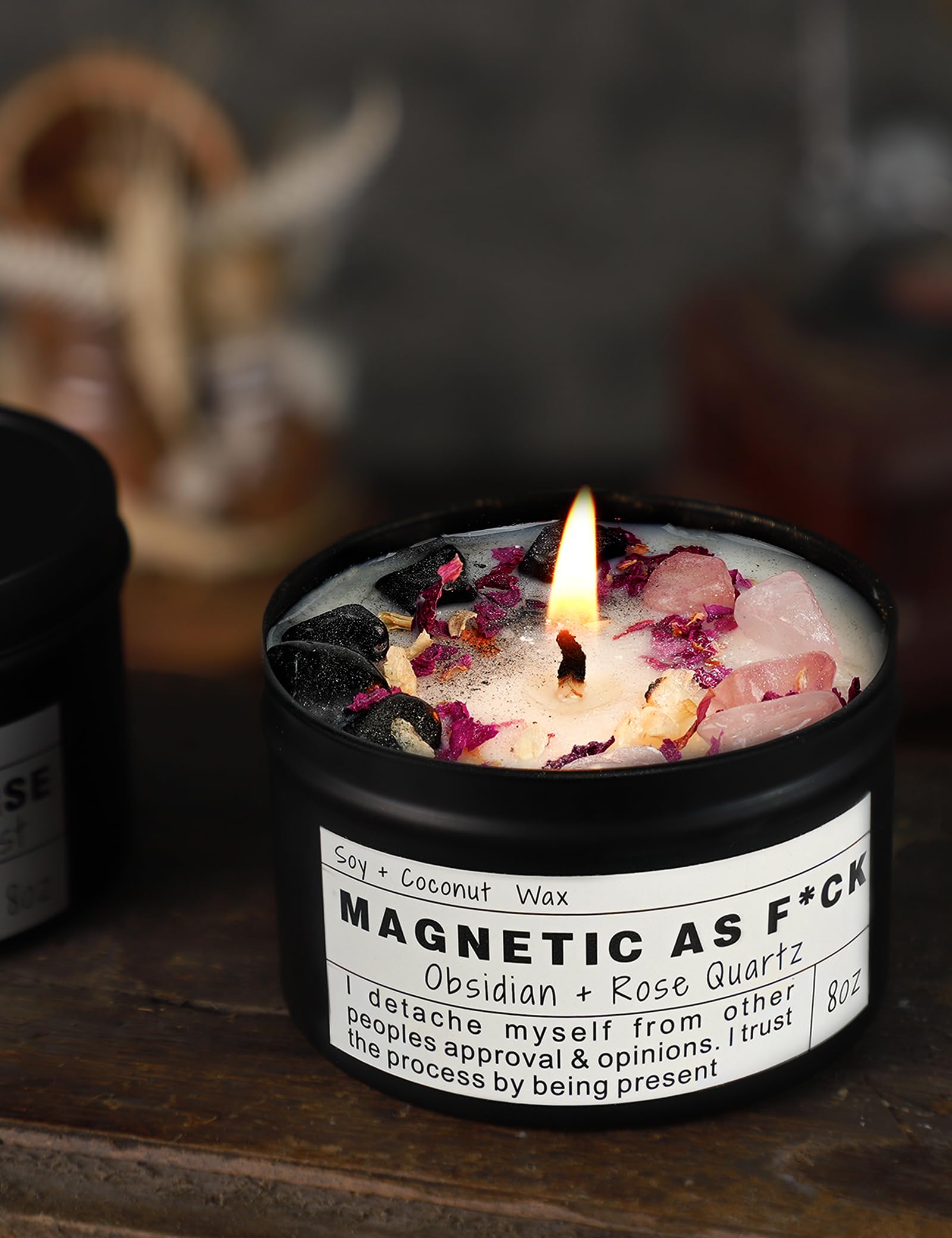 Magnetic As F*ck candle with obsidian and rose quartz crystals burning in a black tin on a wooden surface.