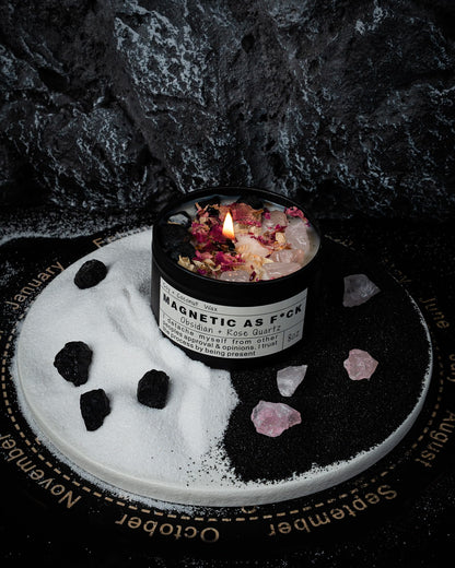 Magnetic As F*ck candle with obsidian and rose quartz crystals displayed on a yin-yang styled sand and stone arrangement against a dark textured background.