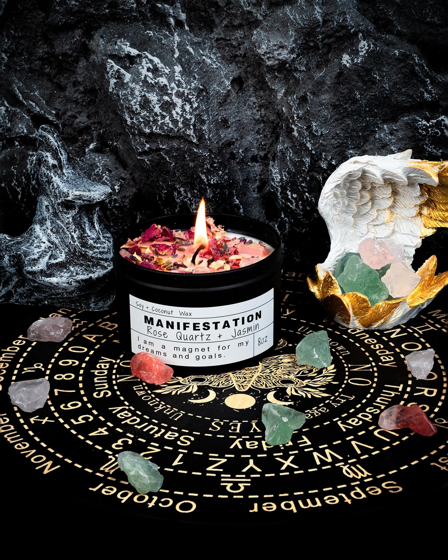 Manifestation candle topped with rose petals and crystals, displayed on a golden mystical wheel with scattered green and pink crystals and an angel wing figurine on a dark rocky background.