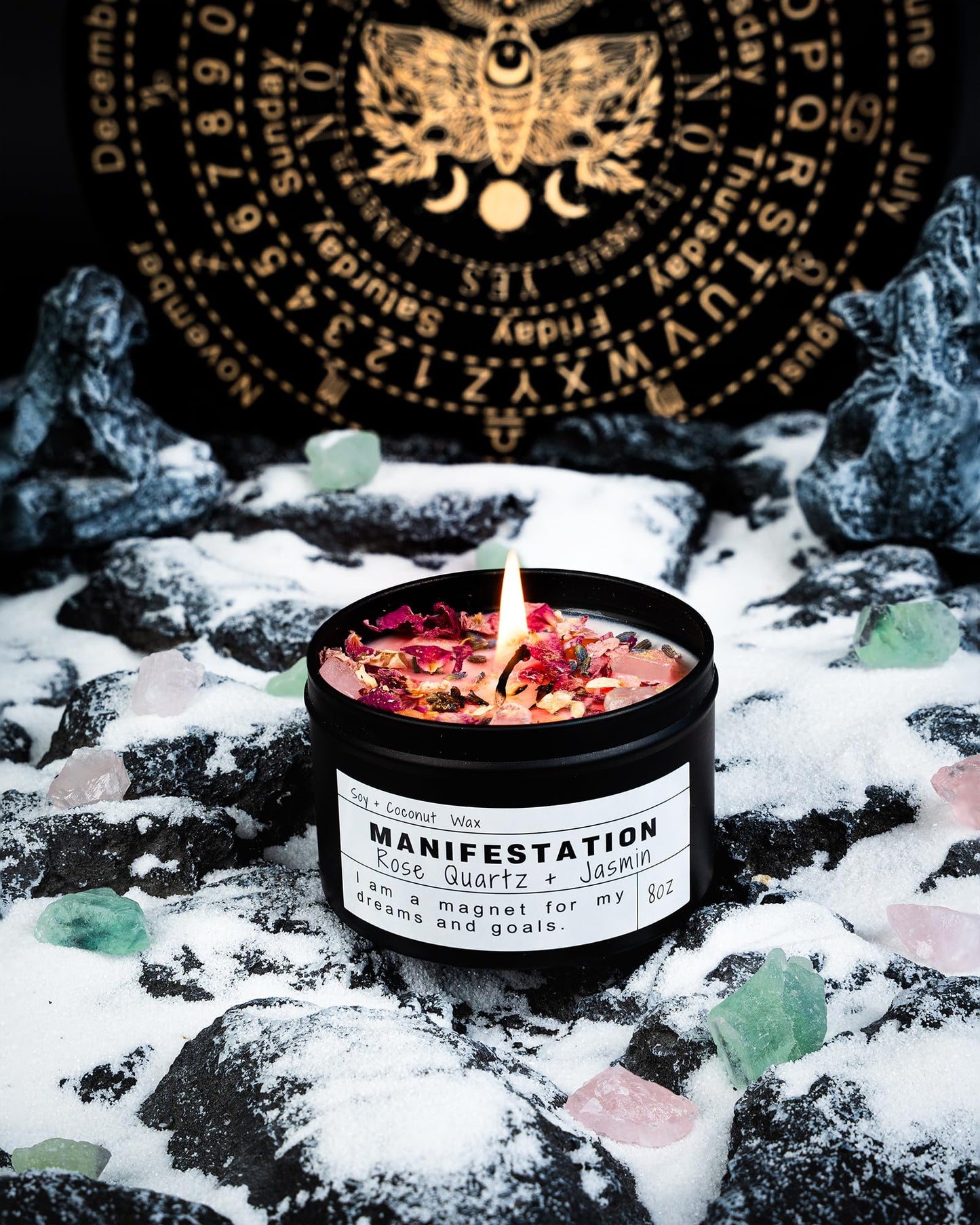 Manifestation candle topped with rose petals and crystals, displayed on snowy black rocks with green and pink crystals and a gold mystical symbol wheel in the background.