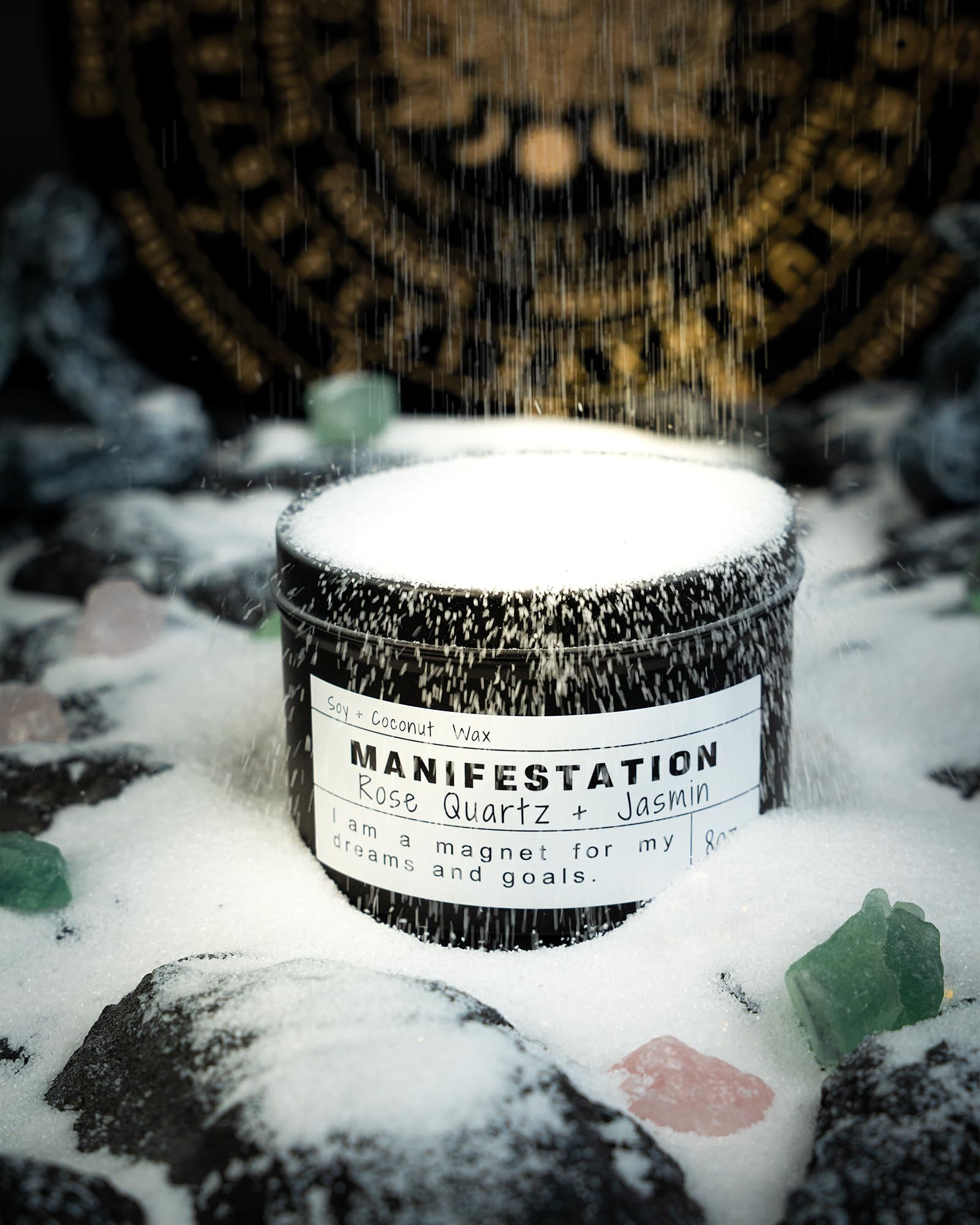 Manifestation candle in a black jar covered with falling snow, surrounded by crystals on a snowy rock surface with a mystical gold symbol wheel in the background