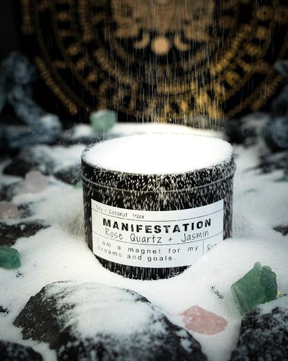 Manifestation candle in a black jar covered with falling snow, surrounded by crystals on a snowy rock surface with a mystical gold symbol wheel in the background